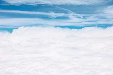 Aerial perspective of vast, fluffy white cloud layer that blankets lower half of the image. Above this dense formation blue sky with some wispy high-altitude clouds and faint contrails is visible