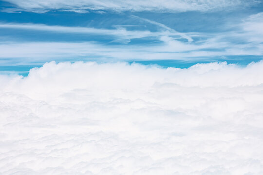 Aerial perspective of vast, fluffy white cloud layer that blankets lower half of the image. Above this dense formation blue sky with some wispy high-altitude clouds and faint contrails is visible