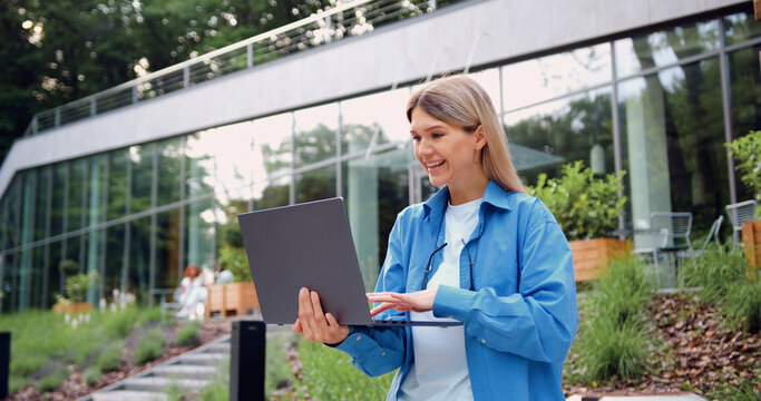 Smiling businesswoman typing on laptop outdoors near modern office building - Powered by Adobe