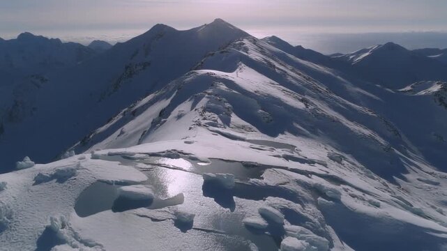 Steep snowy summit casts long shadows across lower ridges, with scattered ice patches reflecting light under a pale winter sky.