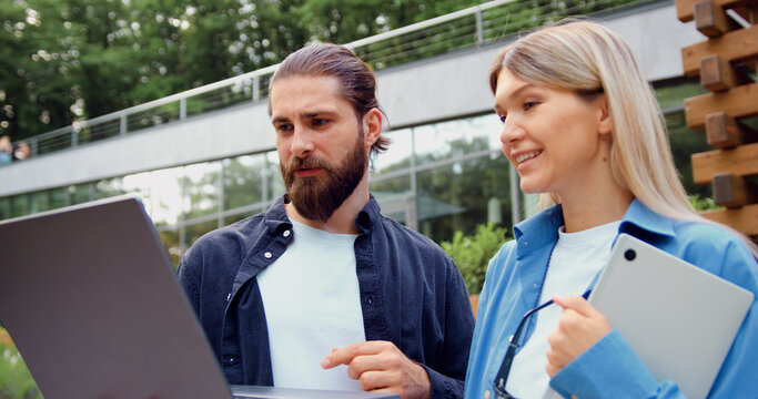 Close-up of two busy, confident business professionals near a modern office building, discussing work tasks with a laptop and tablet, highlighting digital collaboration in an urban environment.