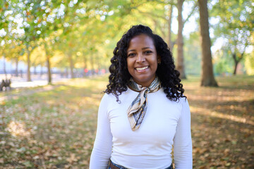 Young African woman smiling at the camera in a tree-lined park during autumn, wearing a scarf and white shirt. Warm backlight and soft bokeh create a relaxed, travel lifestyle mood.