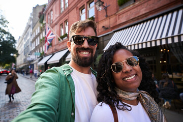 Cheerful mixed-race couple take a selfie looking at the camera on a lively London street, with striped shop awnings and a Union Jack behind them. Sunglasses and scarf add casual travel vibes.