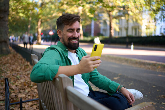 Smiling millennial sits on a park bench in autumn, holding a bright yellow smartphone while checking messages. Urban trees and fallen leaves frame a relaxed, modern lifestyle moment.