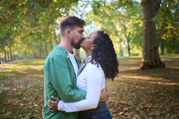 Interracial couple shares a gentle kiss while standing under green and golden trees in a city park. Warm autumn light, fallen leaves and casual outfits convey love, trust and everyday romance.