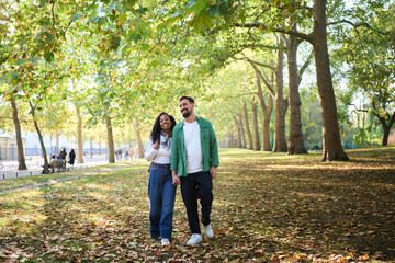Smiling couple strolls hand in hand through a leafy park on a sunny autumn day. Casual outfits, backpack and scarf, relaxed mood and warm light capture a tender moment of travel and city leisure.
