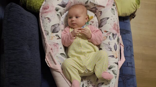 happy, little baby, children's clothes, plays alone with himself, moves his legs cozy cocoon with flower patterns bed. looks camera, tries fingers, chews. Next nipple. Evening. Apartment, house, room