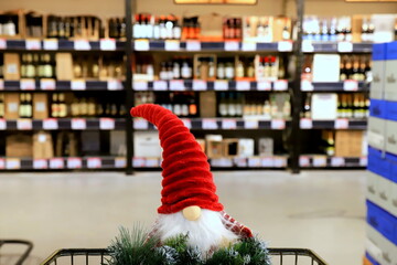 Funny Christmas gnome and wreath on a supermarket trolley. Shopping cart, shelves with bottles of alcohol, food, groceries. Shopping in shop, New Year sale, elite alcohol department for party