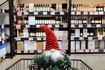 Funny Christmas gnome and wreath on a supermarket trolley. Shopping cart, shelves with bottles of alcohol, food, groceries. Shopping in shop, New Year sale, elite alcohol department for party