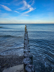 Fototapeta premium Weathered wooden groynes stretch into the gently rippling sea beneath a blue sky streaked with clouds, forming a peaceful coastal scene at the edge of a pebbled beach