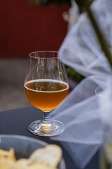 A stemmed glass filled with golden craft beer and a foamy head rests on a dark table, with blurred decorative elements and appetizers nearby.