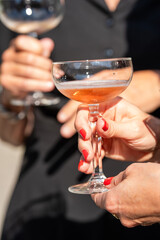 A woman holds a coupe glass filled with sparkling pink beverage. Another glass is blurred in the background, illuminated by bright sunlight.
