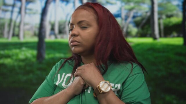 Woman wearing a volunteer shirt clutches her hands at her chest in forest surrounded by trees and greenery; anxiety.