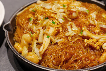 A close-up of savory stir-fried glass noodles (vermicelli) with pieces of seafood (shrimp/squid), cabbage, and egg, tossed in a dark sauce. Chinese or Asian cuisine.