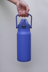 A hand holds a large, textured blue insulated water bottle with a convenient handle and straw lid. The bottle is presented against a neutral light grey background.
