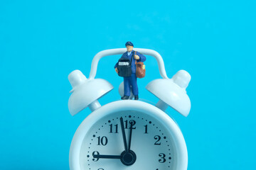 Miniature people toys conceptual photography. Delivery on time service. Postman courier standing above the clock, isolated on blue background.