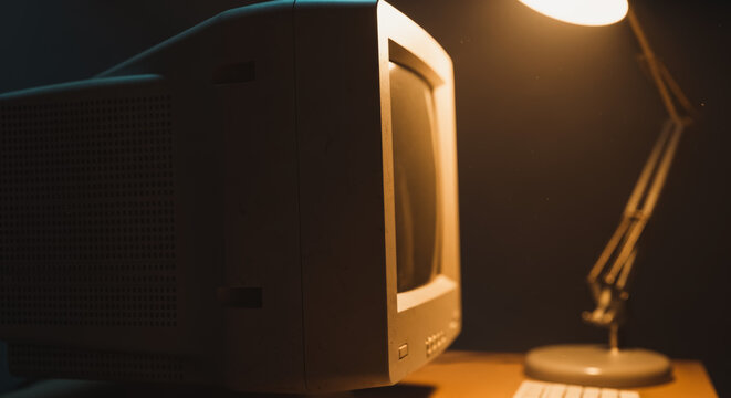An old monitor illuminated by a desk lamp on a simple desk, bathed in warm light, evocative of the past.