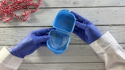 Gloved hands holding an open blue retainer case containing aligners against a wooden background. Candy canes visible, adding a festive touch to the image composition