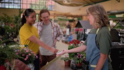 Happy young customer at a flower shop checkout making a contactless payment with a credit card for her purchases, assisted by a smiling female cashier in a bright and lively plant nursery
