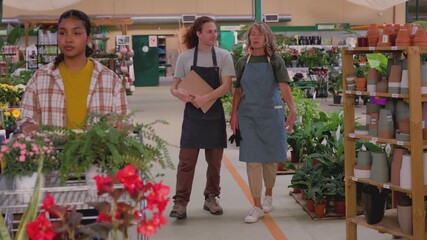 Mature woman manager mentoring a young male employee in a plant nursery, walking through the aisles while a customer pushes a shopping cart full of plants in the foreground - Powered by Adobe
