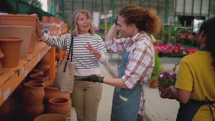 Knowledgeable young male employee in an apron helping a senior woman choose a terracotta plant pot from a shelf in a large, well stocked plant nursery, providing excellent customer service