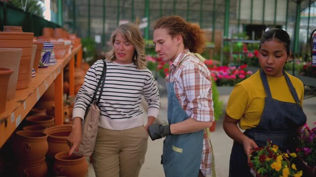 Young male florist in a plaid shirt and apron advising a senior woman on choosing a terracotta pot in a large, bright garden center filled with plants and flowers for her home garden