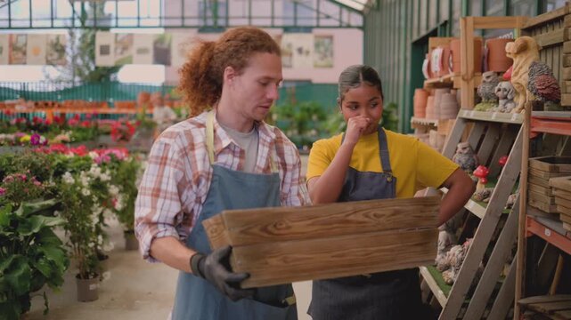 Two diverse young garden center employees collaborating while examining a wooden planter box together, surrounded by colorful flowers and gardening supplies in a vibrant retail nursery setting