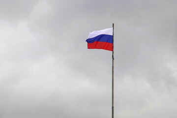 Russian state flag on flagpole waving against dark dense gray clouds in the sky