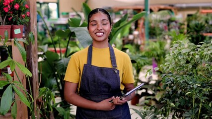 Confident young african american woman working in a flower shop holding a digital tablet. Successful female entrepreneur standing proudly in her small business garden center surrounded by plants - Powered by Adobe