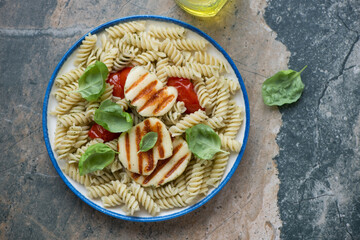Plate of pasta salad with grilled halloumi, tomatoes and basil, horizontal shot on a beige and grey granite background, above view