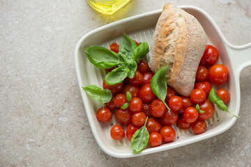 Roasted cherry tomatoes with ciabatta and green basil in a serving tray, horizontal shot on a beige stone background, elevated view