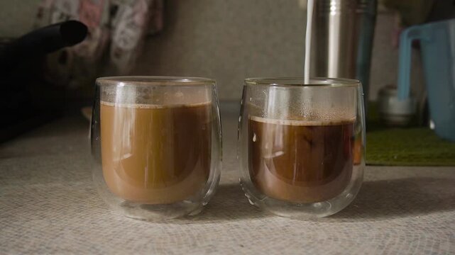 In kitchen, countertop, milk poured into two glasses, cups with double bottom with black coffee. Kitchen utensils background. Morning, bright sunbeams fall from window. Glass shade, steam. Static