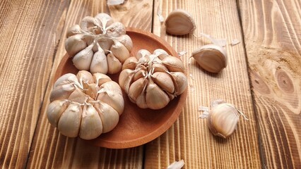 Three whole garlic bulbs sit on a small wooden plate on a rustic wooden surface. The garlic bulbs are intact, with their pale, papery skins visible. Other individual cloves and whole bulbs are scatter