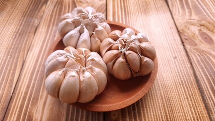 Three whole garlic bulbs sit on a small wooden plate on a rustic wooden surface. The garlic bulbs are intact, with their pale, papery skins visible. Other individual cloves and whole bulbs are scatter