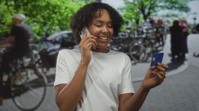 Woman holds smartphone to ear and blue creditcard on sunlit street lined with bicycles and leafy trees; satisfaction.