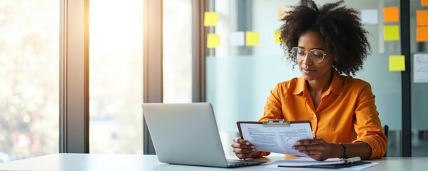 Young African American business woman works at bright modern office desk. Uses laptop computer, reviewing important financial documents on clipboard. Female pro calculates tax reports, provides
