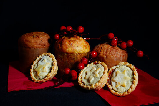 A festive still life of small Panettone cakes and traditional Mince Pies with star tops, decorated with red berries on a dark background. Holiday baking