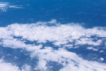 Layer of white cumulus clouds contrasting against blue sky and glimpses of terrain below. Perspective from altitude, looking down over horizontal band of puffy clouds obscuring parts of landscape
