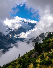 A breathtaking landscape reveals a green, sloped hillside with scattered evergreen trees leading to majestic, cloud-wrapped mountain peaks