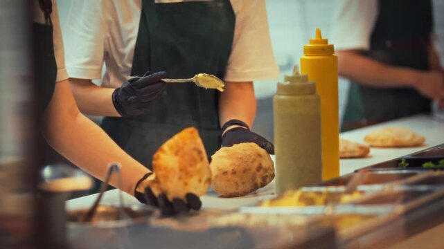 In a fast food kitchen, a chef carefully spreads toppings on freshly baked pita bread. The scene is bustling with energy as food is prepared for eager customers