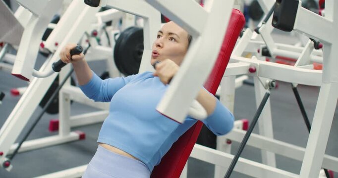Gym workout. Bench press in hammer machine. Slow motion 4k footage of a Caucasian woman trains her chest muscles with focused bench press reps on the hammer machine during an intense gym workout.