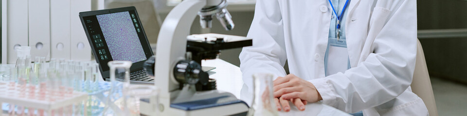 Website banner of woman sitting at laboratory desk operating microscope with glassware and laptop...