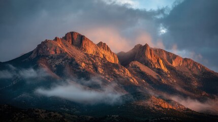 Majestic Mountain Peaks Bathed in Golden Light at Sunset with Cloudy Sky Above and Rolling Fog in the Foreground of a Mountain Range