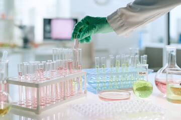 Male hand wearing green glove handling test tube among laboratory glassware with colorful chemical liquids on table in modern medical research laboratory