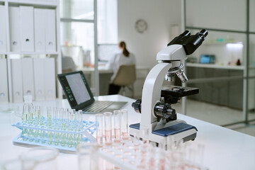 Laboratory table displaying microscope, test tubes with liquid samples, pipette and rack, background showing scientist working at computer in modern medical laboratory