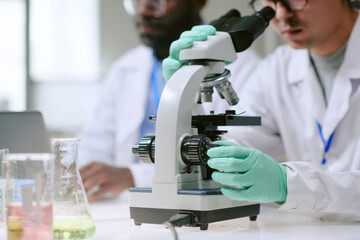 Caucasian man adjusting microscope while young adult Black man observing in laboratory setting, both wearing gloves and lab coats, conducting scientific research