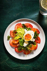 Salad with egg, tomatoes and boiled egg on white plate, healthy meal on dark green background