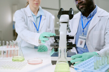 Caucasian woman and Black man working together in laboratory, adjusting microscope and handling glassware, conducting scientific research in medical environment