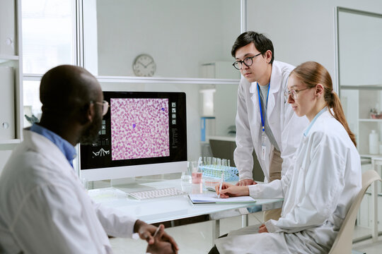 Multiethnic group of scientists analyzing microscopic image on computer monitor in laboratory, discussing medical research results, wearing lab coats