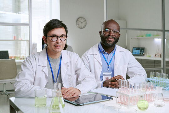Caucasian young adult man and Black middle aged man wearing lab coats sitting at laboratory table smiling, surrounded by glassware, digital tablet, and scientific equipment